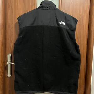 Pu&ograve; includere: Gilet in pile nero con collo alto e sezione superiore in nylon nero. Il gilet presenta il logo bianco "The North Face" sulla parte superiore sinistra del petto. Il gilet &egrave; appeso a un gancio di metallo.
