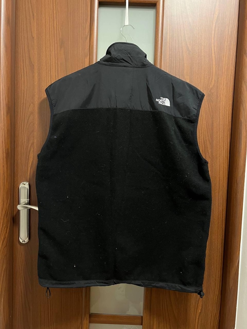 Pu&ograve; includere: Gilet in pile nero con collo alto e sezione superiore in nylon nero. Il gilet presenta il logo bianco "The North Face" sulla parte superiore sinistra del petto. Il gilet &egrave; appeso a un gancio di metallo.
