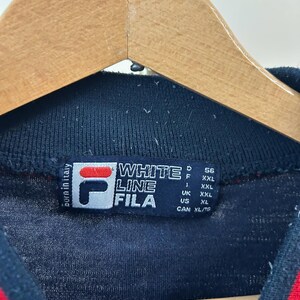 Puede incluir: Una prenda de color azul marino oscuro con cuello acanalado y una etiqueta rectangular que muestra el logotipo de Fila, el texto White Line Fila y Born in Italy. La etiqueta enumera especificaciones de talla para varias regiones: D 56, F XXL, I XXL, UK XXL, US XL y CAN XL/TG.