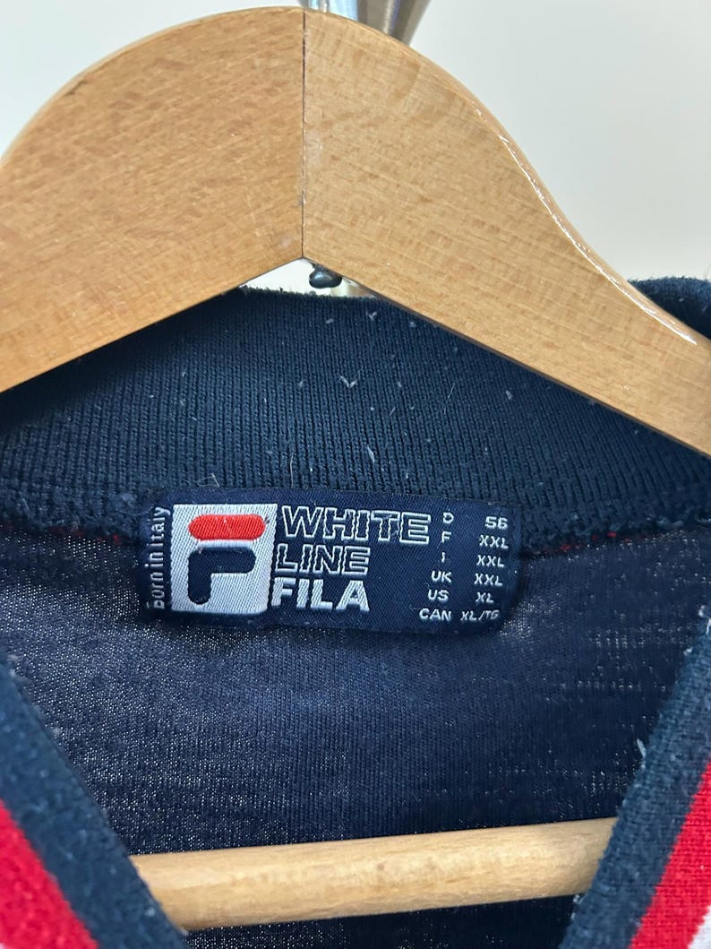 Puede incluir: Una prenda de color azul marino oscuro con cuello acanalado y una etiqueta rectangular que muestra el logotipo de Fila, el texto White Line Fila y Born in Italy. La etiqueta enumera especificaciones de talla para varias regiones: D 56, F XXL, I XXL, UK XXL, US XL y CAN XL/TG.