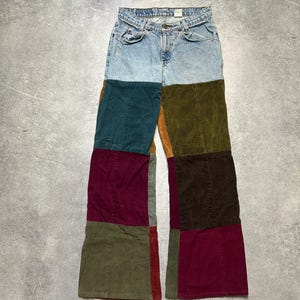 May include: Light wash denim jeans with patchwork corduroy panels in shades of teal, olive green, burgundy, and brown. The jeans have a classic five-pocket design and a button and zipper closure. The patchwork design adds a unique, vintage flair.