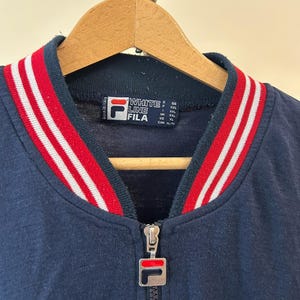 Puede incluir: Una chaqueta azul marino con cremallera y cuello acanalado con rayas rojas y blancas. La etiqueta interior muestra el texto White Line Fila y el tirador de metal de la cremallera lleva el logotipo de Fila. La prenda est&aacute; en una percha de madera.