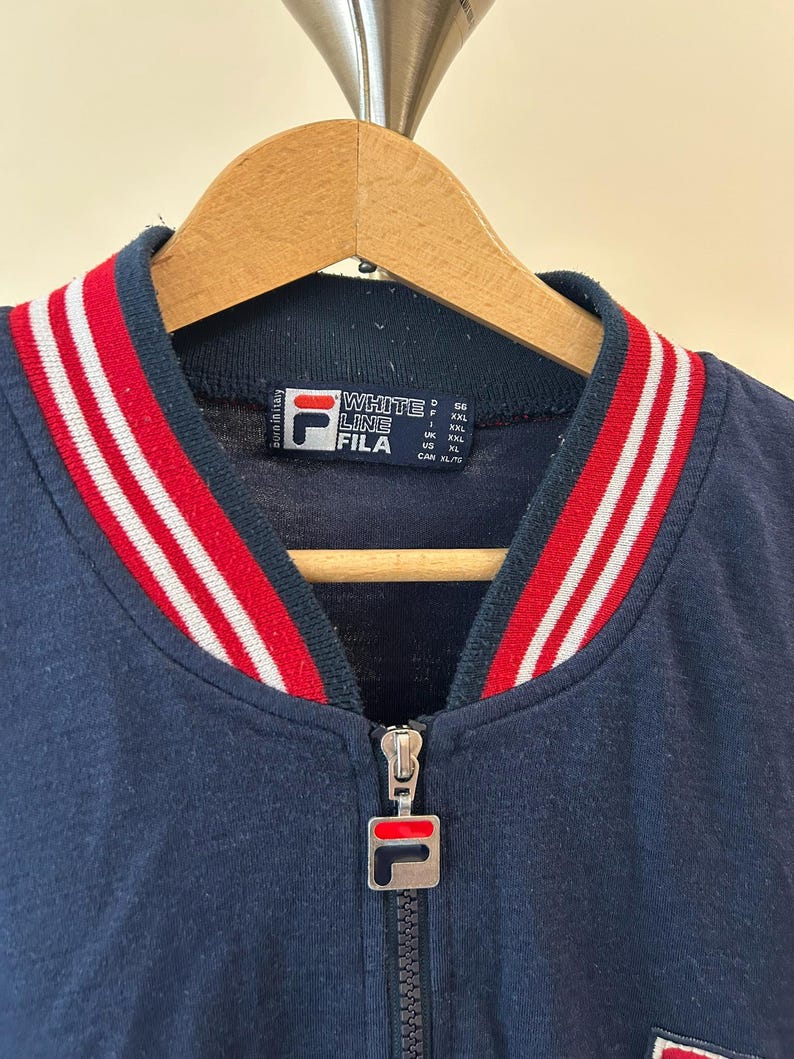Puede incluir: Una chaqueta azul marino con cremallera y cuello acanalado con rayas rojas y blancas. La etiqueta interior muestra el texto White Line Fila y el tirador de metal de la cremallera lleva el logotipo de Fila. La prenda est&aacute; en una percha de madera.