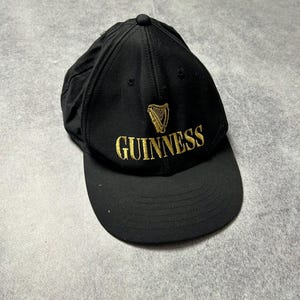Pu&ograve; includere: Cappellino da baseball nero con logo Guinness ricamato in oro. Il logo include un'arpa e la scritta "GUINNESS". Il cappellino ha una visiera curva e un bottone sulla parte superiore.