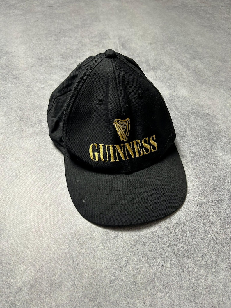 Pu&ograve; includere: Cappellino da baseball nero con logo Guinness ricamato in oro. Il logo include un'arpa e la scritta "GUINNESS". Il cappellino ha una visiera curva e un bottone sulla parte superiore.