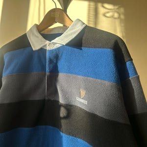 May include: A blue, black, and gray striped fleece rugby shirt with a white collar and the Guinness logo. The shirt is hanging on a wooden hanger.