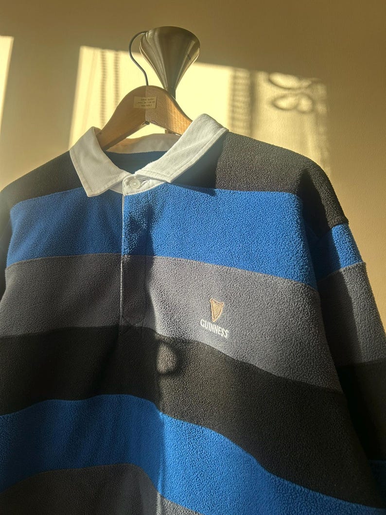 May include: A blue, black, and gray striped fleece rugby shirt with a white collar and the Guinness logo. The shirt is hanging on a wooden hanger.