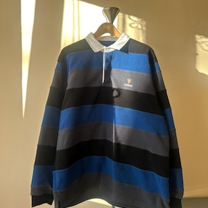 May include: A long-sleeved rugby shirt with horizontal stripes in black, blue, and gray. The shirt has a white collar and a small Guinness logo on the chest. The shirt is hanging on a wooden hanger.