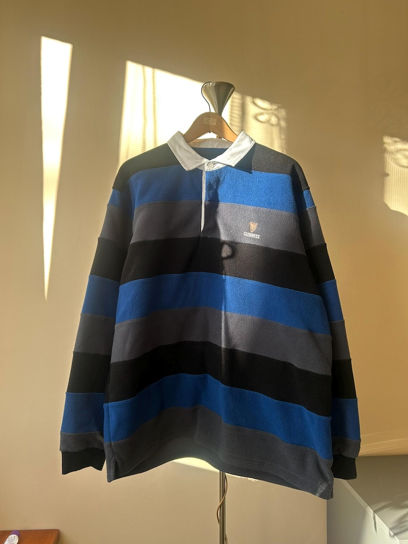 May include: A long-sleeved rugby shirt with horizontal stripes in black, blue, and gray. The shirt has a white collar and a small Guinness logo on the chest. The shirt is hanging on a wooden hanger.