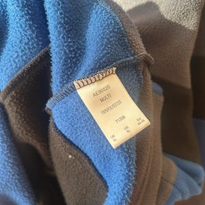 May include: Close-up of a blue, gray, and black fleece garment. The tag reads "AE90820 MULTI 100% POLYESTER" with size information. The fleece has a soft, textured appearance.