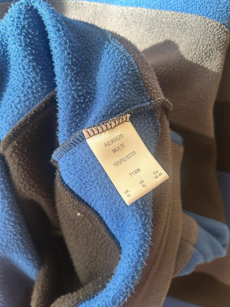 May include: Close-up of a blue, gray, and black fleece garment. The tag reads "AE90820 MULTI 100% POLYESTER" with size information. The fleece has a soft, textured appearance.