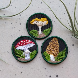 Puede incluir: Tres parches bordados para planchar con diferentes tipos de setas. Los parches son circulares y tienen un borde verde. El primer parche presenta una seta amarilla con manchas blancas. El segundo parche presenta una seta roja con manchas blancas. El tercer parche presenta una seta marrón con una superficie texturizada.
