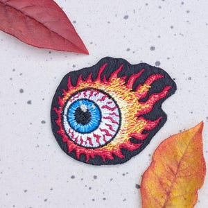 May include: Embroidered patch featuring a flaming eyeball design. The eye has a blue iris, red veins, and a black outline. The flames are yellow and red. The patch is on a black background. The patch is surrounded by fall leaves.