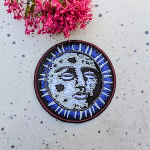 May include: Embroidered iron-on patch featuring a white and gray moon face with a blue and white starburst design on a black background.