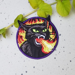 Pode incluir: Etiqueta bordada de um gato preto com olhos verdes que cospe fogo. O gato está rodeado de chamas em tons de vermelho, laranja e amarelo. A etiqueta tem uma borda roxa.