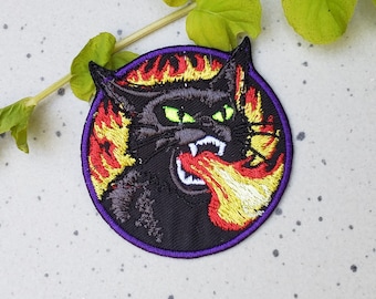 May include: Embroidered patch of a black cat with green eyes breathing fire. The cat is surrounded by flames in shades of red, orange, and yellow. The patch has a purple border.