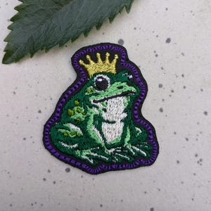 May include: Embroidered patch featuring a green frog wearing a gold crown. The frog has a white chest and is outlined in black with a purple border. The patch is likely for clothing or accessories.