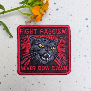 May include: Embroidered patch with a black cat design and the words "FIGHT FASCISM" and "NEVER BOW DOWN" in red lettering. The patch has a black background with a red border and red rays emanating from the cat.