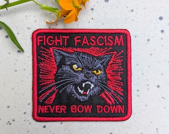 May include: Embroidered patch with a black cat design and the words "FIGHT FASCISM" and "NEVER BOW DOWN" in red lettering. The patch has a black background with a red border and red rays emanating from the cat.