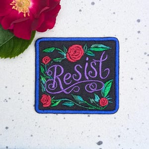 May include: Square embroidered patch with the word "Resist" in purple script, surrounded by red roses and green leaves on a black background. The patch has a blue border. A red rose is in the upper left corner.
