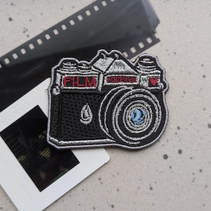 May include: Black and silver embroidered iron-on patch of a vintage camera with the words "FILM FOREVER" and a red heart.