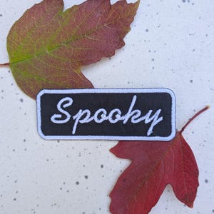 May include: A black and white embroidered patch with the word "Spooky" in white cursive lettering. The patch is surrounded by a white border and is sitting on a white surface with two red leaves.