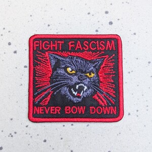 May include: Embroidered patch with a black cat illustration, red accents, and the text "FIGHT FASCISM NEVER BOW DOWN." The cat has yellow eyes and an angry expression. The patch is square with a red border.
