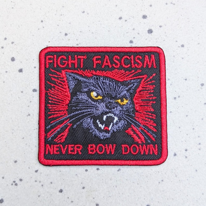 May include: Embroidered patch with a black cat illustration, red accents, and the text "FIGHT FASCISM NEVER BOW DOWN." The cat has yellow eyes and an angry expression. The patch is square with a red border.