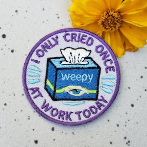 May include: Purple and white embroidered patch with a blue tissue box that says "weepy" and an eye on the front. The text around the tissue box says "I only cried once at work today".