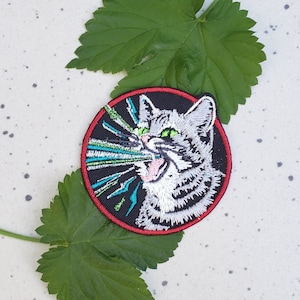 May include: A round embroidered patch featuring a white cat with green eyes, a black background, and a red border. The cat is depicted with its mouth open, as if it is yelling or screaming. The cat's eyes are glowing green, and there are green and blue lines radiating from its mouth.