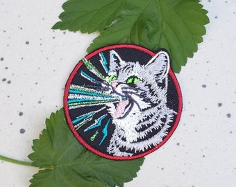 May include: A round embroidered patch featuring a white cat with green eyes, a black background, and a red border. The cat is depicted with its mouth open, as if it is yelling or screaming. The cat's eyes are glowing green, and there are green and blue lines radiating from its mouth.
