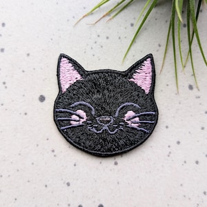 May include: Black embroidered cat face patch with pink ears and a smiling expression.