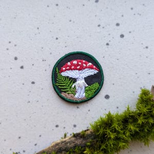 May include: A red and white mushroom with white spots, embroidered on a green and black fabric patch. The mushroom is surrounded by green leaves.