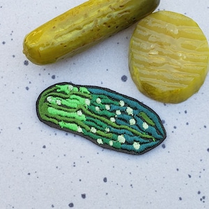 May include: An embroidered patch of a pickle, featuring green and white stitching. The patch is displayed on a white background with black speckles.