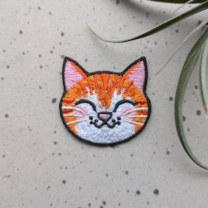May include: Embroidered iron-on patch of a smiling orange tabby cat with white fur around the eyes and mouth.