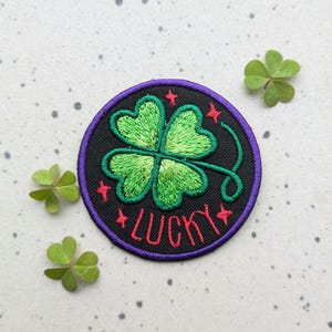 Puede incluir: Un parche bordado negro y morado con un trébol de cuatro hojas verde y la palabra "LUCKY" en rojo.