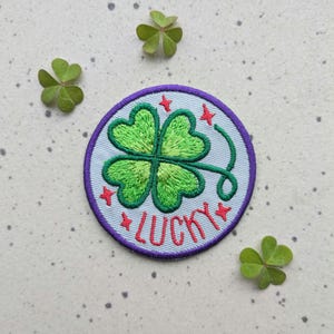Puede incluir: Parche bordado con un trébol verde de cuatro hojas y la palabra "LUCKY" en rojo. El parche tiene un fondo azul claro con acentos de estrellas rojas y un borde morado. Tres tréboles verdes están dispersos alrededor del parche.