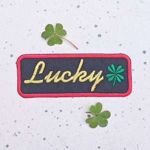 May include: A black and red embroidered patch with the word "Lucky" in gold lettering and a green four-leaf clover.