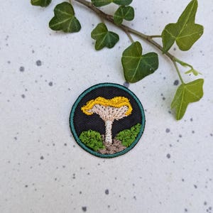 May include: A round embroidered patch with a yellow and white mushroom on a dark green background. The mushroom has a yellow cap and a white stem. The background is a dark green with a light green border.