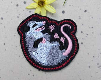 May include: A black embroidered patch with a red border featuring a gray and white opossum with a pink tail curled up in a ball.