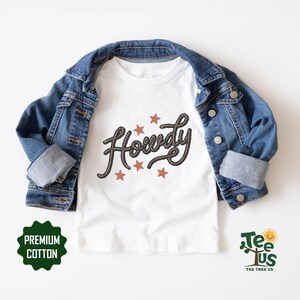 May include: White t-shirt with the word "Howdy" written in a rope-like font with brown stars. The shirt is on a white background and is folded in half. The shirt is made of premium cotton.