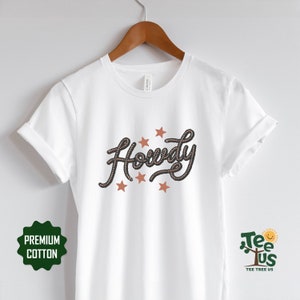 May include: White t-shirt with the word "Howdy" written in a rope-like font with brown stars around it. The shirt is on a hanger and has a tag that says "Canvas" on it.