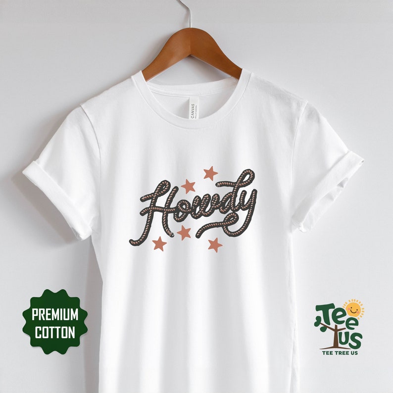 May include: White t-shirt with the word "Howdy" written in a rope-like font with brown stars around it. The shirt is on a hanger and has a tag that says "Canvas" on it.