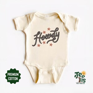 May include: A cream-coloured baby onesie with short sleeves and a snap closure. The onesie has a black and brown rope-style graphic that says "Howdy" with five red stars around it.