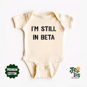 May include: A cream-coloured baby onesie featuring the text "I'M STILL IN BETA" in bold black lettering. The onesie has short sleeves and a snap closure. A green "Premium Cotton" badge and a logo are visible.