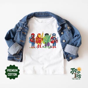 May include: A white t-shirt featuring a graphic of four cartoon superheroes carrying school supplies, layered under a blue denim jacket. The graphic includes Spider-Man, Iron Man, Hulk, and Captain America with backpacks, pencils, and books.