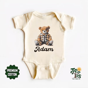 May include: A cream-colored baby onesie with a brown teddy bear wearing a plaid shirt. The onesie has the name "Adam" printed in brown cursive lettering.
