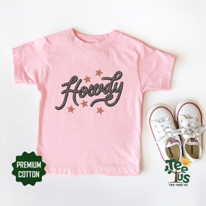 May include: Pink short-sleeved t-shirt with the word "Howdy" written in a rope-like font with brown stars. The shirt is made of premium cotton.