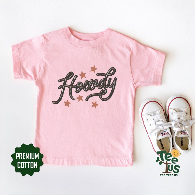 May include: Pink short-sleeved t-shirt with the word "Howdy" written in a rope-like font with brown stars. The shirt is made of premium cotton.