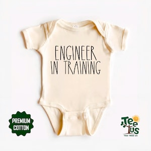 May include: Cream-colored baby onesie with the text "ENGINEER IN TRAINING" in black. The onesie has short sleeves and a round neckline. A green and white "PREMIUM COTTON" badge is on the lower left. The Tee Tree Us logo is on the lower right.